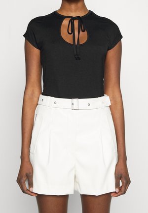 Vrouw draagt een zwarte top met korte mouwen, een keyhole-halslijn en een strik, gecombineerd met witte shorts met hoge taille, riem en ritssluitingen in de zakken.