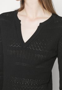 Femme portant un haut noir en maille à col en V avec des détails ajourés à motifs.