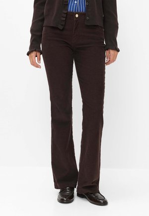 Person iført mørkebrune kordbuks, sorte loafers, en sort cardigan og en blåstribet skjorte, stående mod en hvid baggrund.
