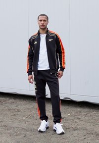 Survêtements de sport noir avec des rayures orange-jaune, comprenant une veste zippée et un pantalon à taille élastique, associés à des baskets blanches.