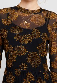 Svart genomskinlig långärmad klänning med orange blommönster, lager över en svart camisole, tillsammans med två delikata guldhalsband.