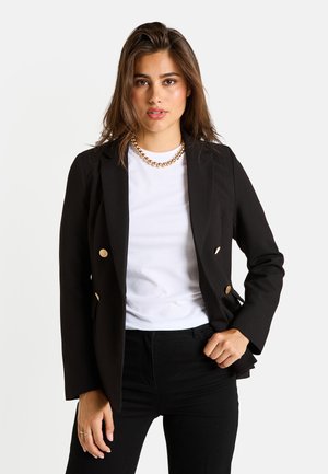 Mujer joven con un blazer negro de doble botonadura, camisa blanca, pantalones negros y un collar de cuentas doradas, sobre un fondo liso.
