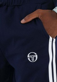 Marine sportbroek met witte zijstrepen, voorzien van een geborduurd cirkelvormig logo op het onderbeen. De stof heeft een gestructureerde afwerking.