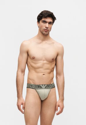 Homme musclé aux cheveux courts et foncés portant un slip gris clair Emporio Armani sur un fond blanc uni.