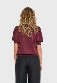 Mujer con cabello rubio largo y rizado, vistiendo una blusa burdeos de mangas abullonadas y pantalones negros, vista desde atrás contra un fondo simple.