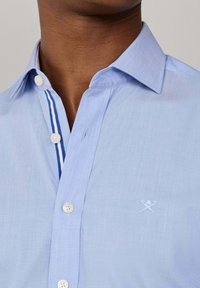 Detailní záběr osoby nosící světle modrou košili na knoflíky s bílými knoflíky a modře proužkovaným vnitřním lemem knoflíkové légy, s malým vyšívaným logem na hrudi.