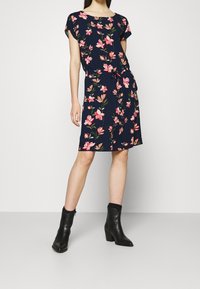 Marinblå klänning med blommönster i rosa och grönt, korta ärmar, rund halsringning och åtstramad midja. Buren med svarta klackstövletter.