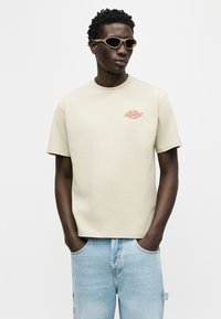 Άνδρας που φοράει μπεζ κοντομάνικο μπλουζάκι με μικρό κόκκινο λογότυπο στο στήθος, ανοιχτό μπλε τζιν και μπεζ γυαλιά ηλίου, με τα χέρια στις τσέπες.