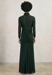 Pikk, keha külge liibuv tume roheline kleit kõrge kraega, pikkade varrukatega ja volditud detailidega seljal. Valmistatud siledast, venivast kangast.