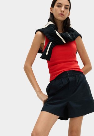 Kvinde iført rød ærmeløs top, sorte shorts og sort sweater hængende over skuldrene, står med hænderne på hofterne.