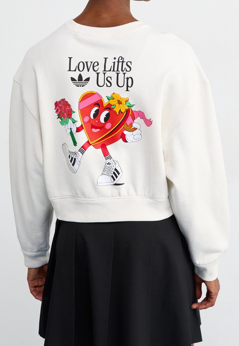 Cream sweatshirt featuring a playful heart graphic, "Love Lifts Us Up," with floral accents and a black skirt. Black and white Adidas sneakers visible.