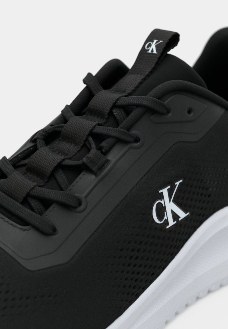 Chaussure de sport noire avec des lacets noirs, semelle blanche et logo "cK" blanc sur le côté et la languette.