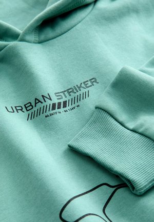 Minttuvihreä huppari, jossa on ribbilehdet. Tässä on musta teksti "URBAN STRIKER" ja koordinaatit. Sileä pinta, rento muotoilu.