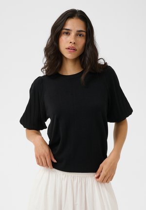 Femme aux cheveux bruns ondulés, portant un haut noir à manches courtes et une jupe blanche, debout devant un fond clair uni.
