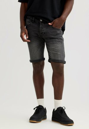 Hombre con pantalones cortos de mezclilla gris oscuro remangados, zapatillas negras, calcetines blancos y una camisa negra con una mano en el bolsillo.