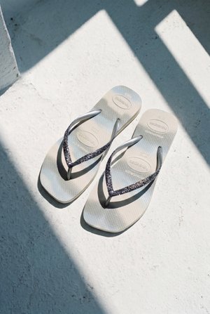 Egy pár világos színű Havaianas flip-flop csillogó sötét pántokkal, napsütötte, texturált fehér padlón árnyékokkal.