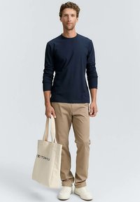 Temno modra majica z dolgimi rokavi, bež hlače in bele superge. Drži svetlo barvno torbo z črnim logotipom. Preprost, priložnostni slog.