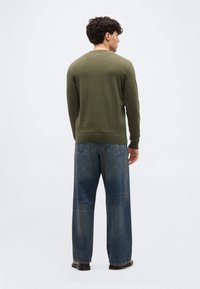 Homme debout de dos, portant une chemise manches longues vert olive, un jean bleu ample et des chaussures noires, devant un fond uni.