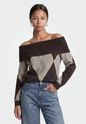 Donna con capelli scuri che indossa orecchini a cerchio, un maglione a rombi marrone e grigio con scollo a barca e jeans blu, con una mano in tasca.