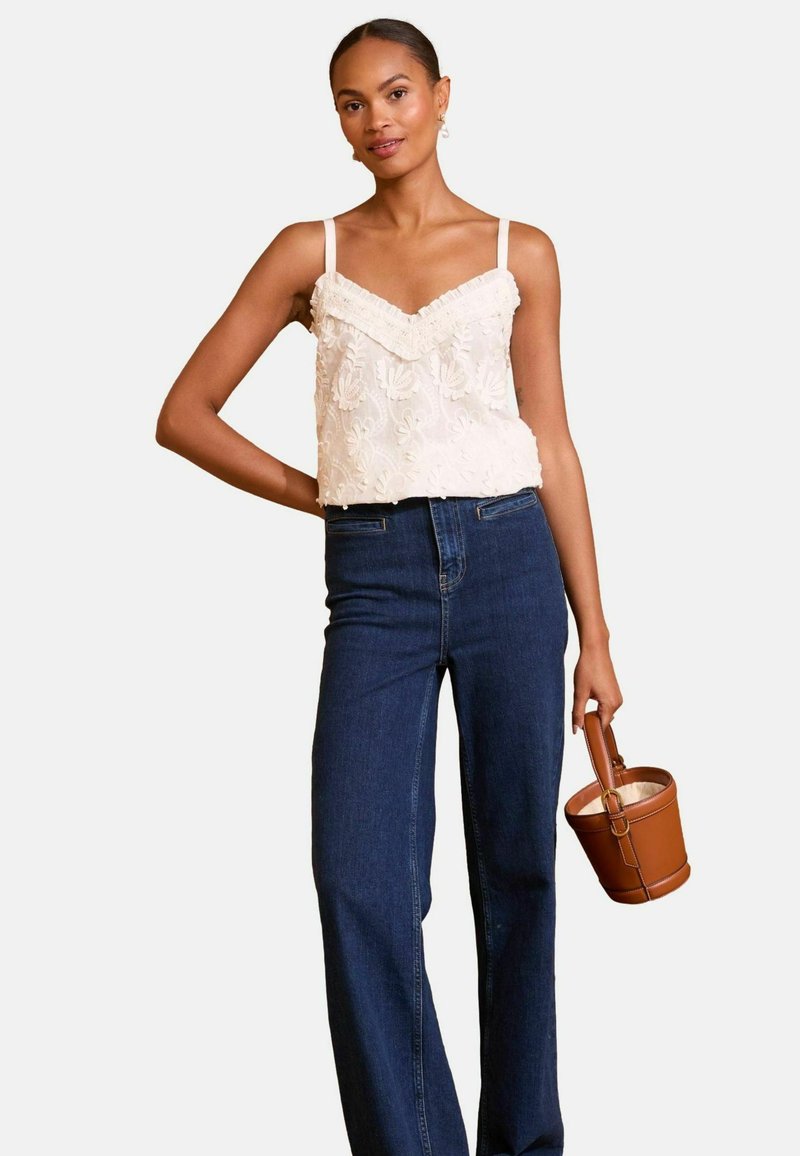 Kvinde iført en broderet hvid ærmeløs top, højtaljede blå jeans, holder en brun læder bucket bag mod en ensfarvet baggrund.