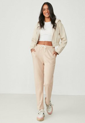 Beige Cropped-Hoodie mit Kordelzug, kombiniert mit hellrosa Jogginghosen mit elastischem Bund und Seitentaschen. Getragen mit mehrfarbigen Sneakern.