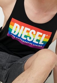 Svart linne med en flerfärgad regnbågsdesign som visar "DIESEL FOR SUCCESSFUL LIVING" i fet vita bokstäver. Grå shorts synliga.