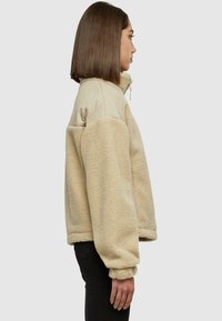 Žena u bež flis jakni s najlonskim ramenim panelima stoji u profilu na svijetlosivoj pozadini.