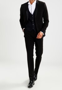Schwarzer Anzug mit schmaler Passform, Satin-Revers und strukturiertem Weste. Kombiniert mit schwarzen Dressschuhen und einem weißen Hemd.
