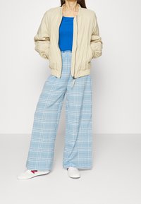 Giacca bomber beige, canotta blu, pantaloni a palloncino a quadri blu chiaro. Sneakers bianche con accenti rossi.