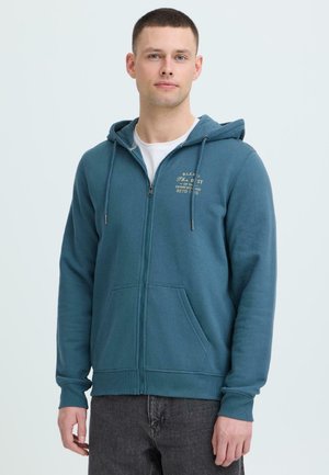 Mężczyzna ubrany w turkusową bluzę z zamkiem błyskawicznym i kieszeniami z przodu, z nadrukiem na lewej piersi, w połączeniu z ciemnoszarymi jeansami na jednolitym tle.