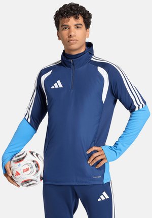 Młody mężczyzna w niebieskiej odzieży sportowej Adidas trzyma białą i czarną piłkę do piłki nożnej Adidas Tiro, jedną ręką opierając się na biodrze.