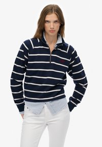 Femme portant un pull à col zippé bleu marine et blanc à rayures avec une broderie "S.DRY NYC", superposé sur une chemise à rayures bleues et un pantalon blanc.