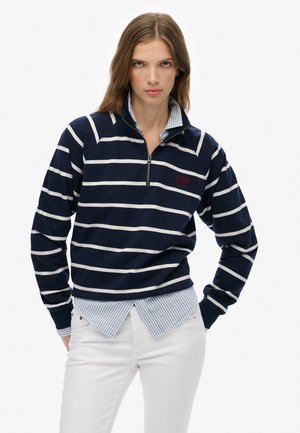 Femme portant un pull à col zippé bleu marine et blanc à rayures avec une broderie "S.DRY NYC", superposé sur une chemise à rayures bleues et un pantalon blanc.