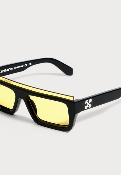 Lunettes de soleil rectangulaires noires avec des verres jaunes et une bande dorée sur la monture supérieure, avec un logo blanc sur la branche.