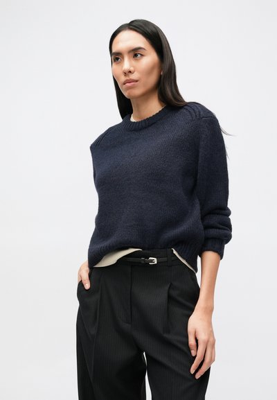 Sieviete, kura valkā tumši zilu džemperi virs baltas blūzes, melnas bikses ar smalkām svītrām un melnu jostu, stāv pret vienkāršu gaišu fonu.