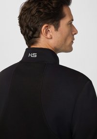 Muškarac u crnoj majici s visokim ovratnikom na kojem se nalaze bijela slova "HS", prikazan s leđa i sa strane na jednoličnoj svijetloj pozadini.