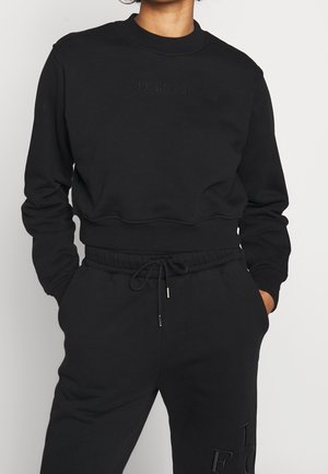 Osoba nosząca czarną bluzę z dekoltem typu crewneck z wytłoczonym napisem "Fiorucci" na piersi oraz dopasowane czarne spodnie dresowe ze sznurkiem w talii i wytłoczonymi literami.