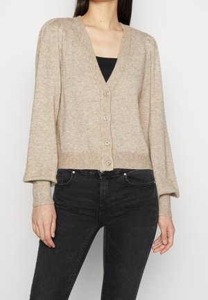 Gilet en tricot beige avec un col en V, cinq boutons, des poignets côtelés et des manches froncées. Associé à un jean noir ajusté.