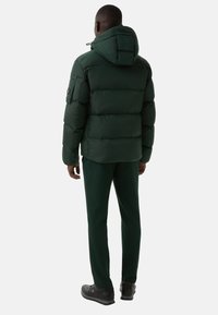 Dark green puffer jacket with hood, horizontal stitching, and logo on the back. Paired with matching green pants and black shoes.