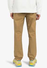 Khakifärgade bomullsbukser med rak benpassform, i kombination med beige och gula sneakers med blå detaljer. Enkelt design, inga synliga mönster.