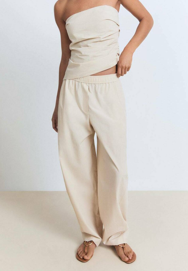 En person står iført en stropløs beige top og løstsiddende beige bukser, med bare fødder synlige. Baggrunden er lys og neutral.