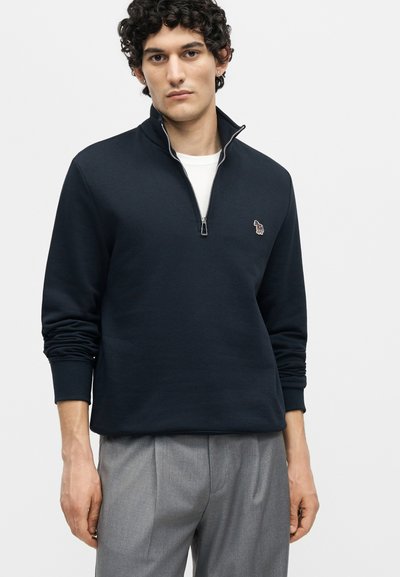 Άνδρας με σγουρά μαλλιά, που φοράει ένα μπλε ναυτικό πουλόβερ με φερμουάρ μέχρι το μέσο πάνω από λευκό πουκάμισο και γκρι παντελόνια, στέκεται μπροστά σε απλό φόντο.