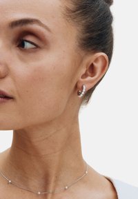 Zilveren oorring in een cirkelvorm met kleine, heldere edelstenen; dunne zilveren ketting met gelijkmatig verdeelde parels; neutrale huidtint en haar naar achteren gebonden.