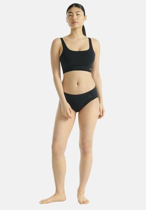 Jonge vrouw staat blootsvoets, gekleed in een zwarte sportbeha en bijpassende slip, tegen een effen lichte achtergrond.