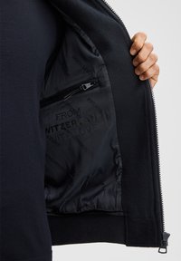 Black zip-up jacket with a smooth inner lining, featuring a zippered pocket and printed text reading "FROM SWITZERLAND WITH LOVE."