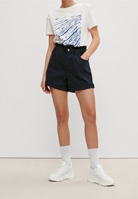 T-shirt blanche avec un motif graphique bleu, shorts taille haute bleu marine, et baskets blanches. Les shorts présentent des revers et des poches latérales.