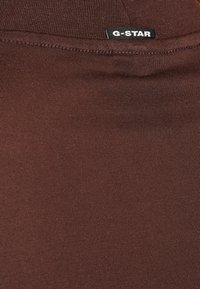 Brun bomullstyg med en slät textur och en G-Star-logotyp sydd upptill. Materialet är elastiskt och kroppsnära.