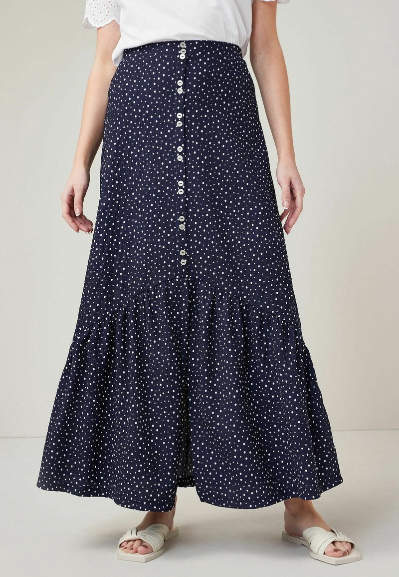 Femme portant une jupe bleu marine longueur cheville avec des pois blancs et des boutons devant, associée à des sandales blanches et un haut blanc.