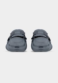 Σκούρα γκρι σανδάλια με χοντρές, σκαλιστές σόλες και ρυθμιζόμενα λουριά, εμφανίζονται από πίσω σε απλό ανοιχτό φόντο.