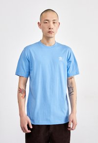 Camiseta de algodón de color azul claro con cuello redondo y mangas cortas. Presenta un pequeño logo blanco de Adidas en el lado izquierdo del pecho. Diseño simple.